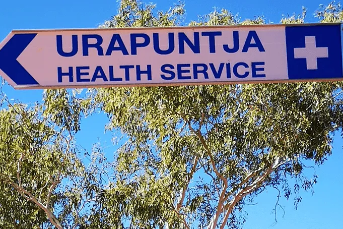 Road sign against a blue sky: Urapuntja Health Service, with an arrow pointing left